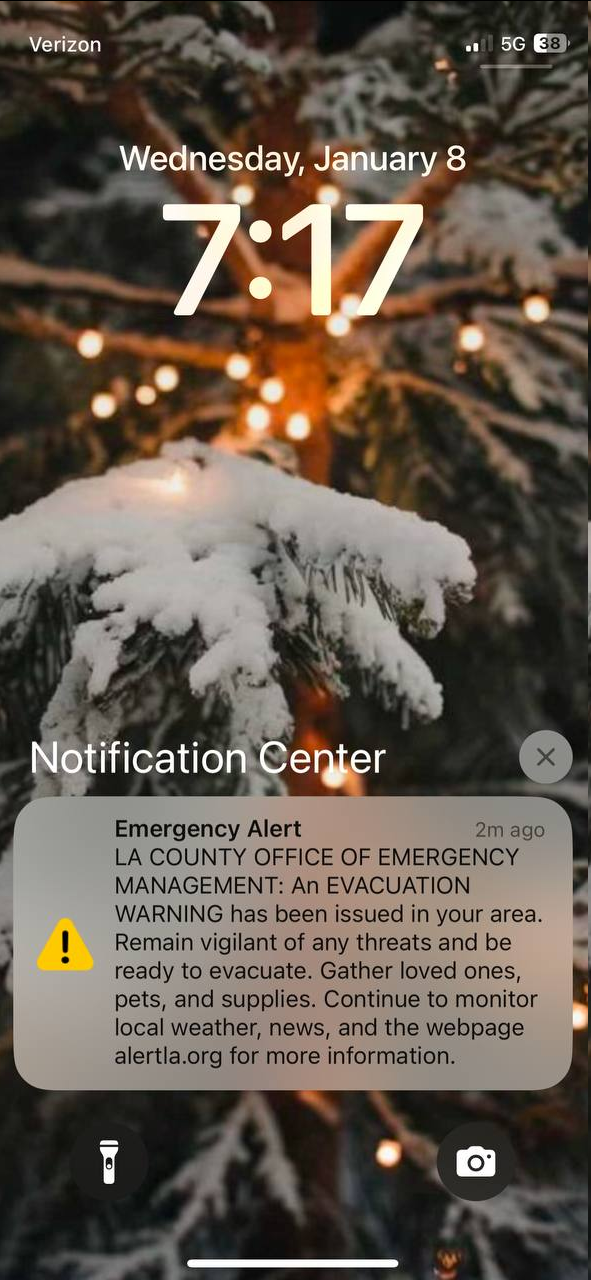 A Wednesday, January 8, 7:17am screenshot of an Emergency Alert on a Christmas-y home screen sent 2 minutes ago that reads: Emergency Alert. LA COUNTY OFFICE OF EMERGENCY MANAGEMENT: An EVACUATION WARNING has been issued in your area. Remain vigilant of any threats and be ready to evacuate. Gather loved ones, pets, and supplies. Continue to monitor local weather, news, and the webpage alertla.org for more information."
