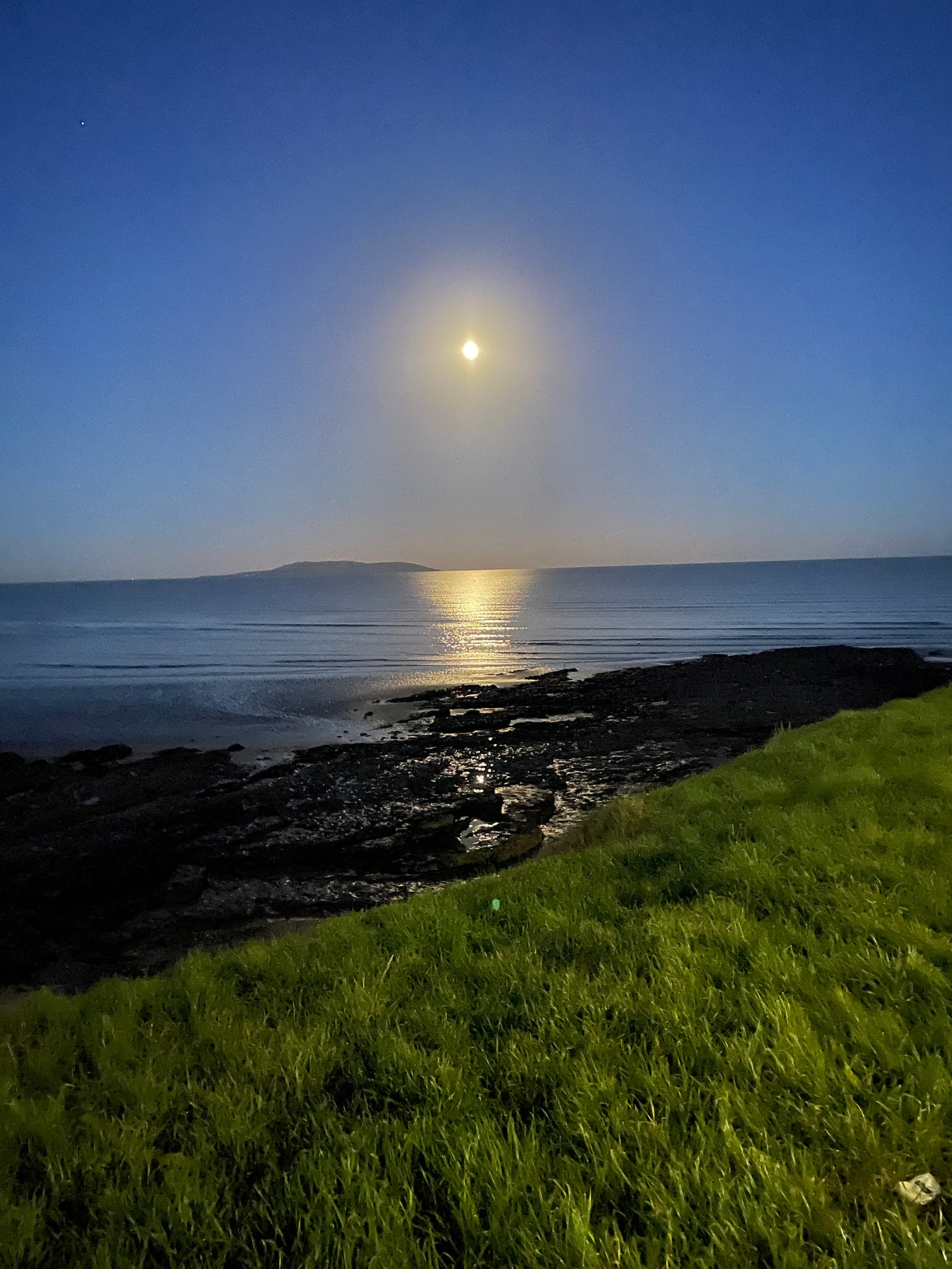 A super bright, probably full moon reflecting brightly across the Irish Sea. Grass and rocks are in the foreground.