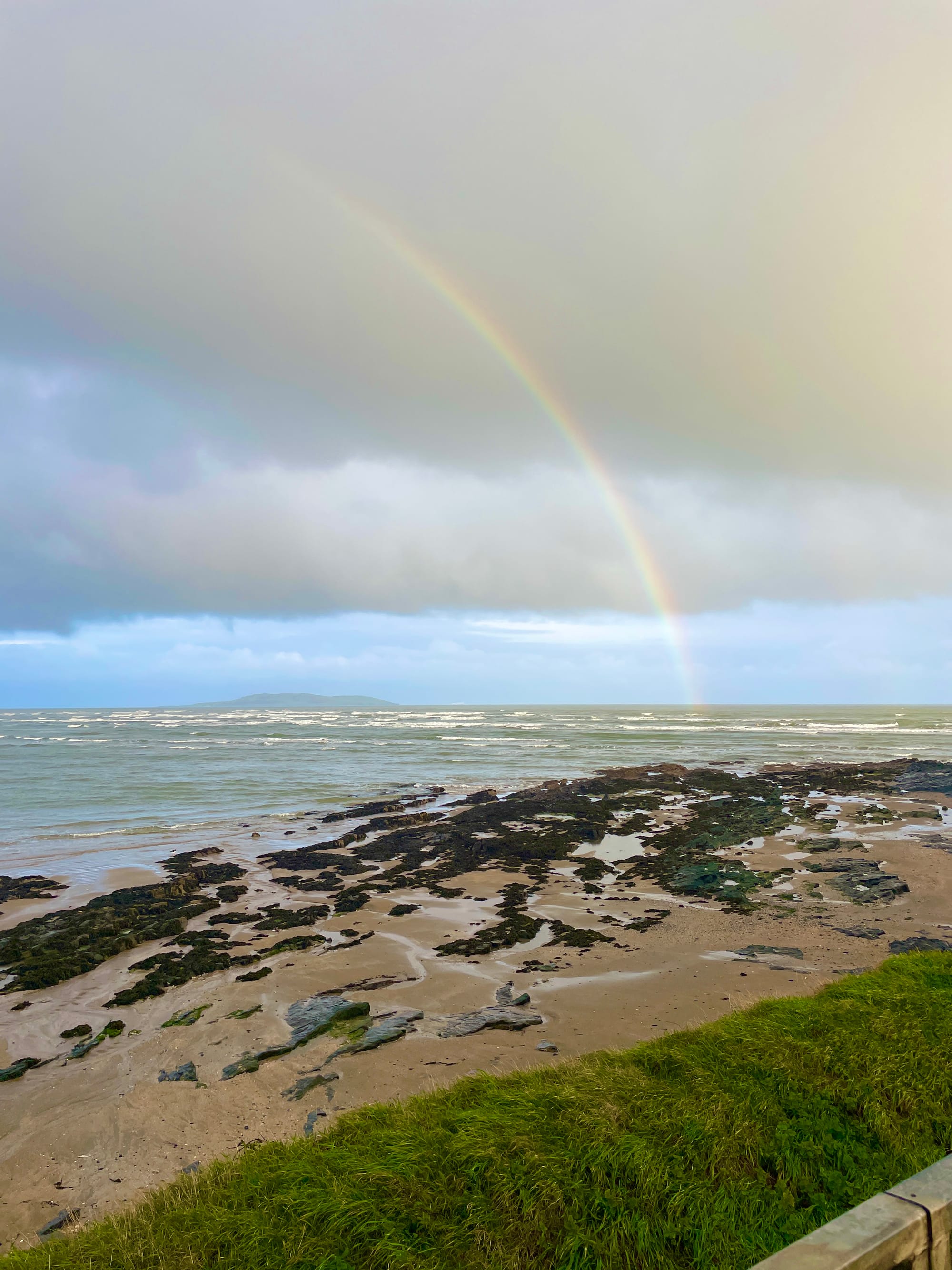 rainbow amongst the clouds, over a rocky beach and a choppy Irish Sea